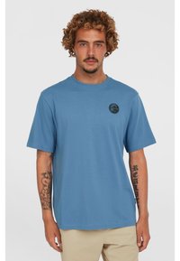 Light blue cotton t-shirt with short sleeves, round neck, and a circular black logo on the left chest. Features a relaxed fit.