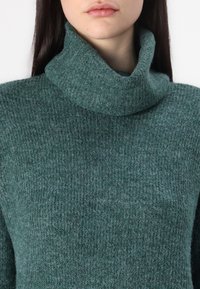 Pull en laine bleu sarcelle avec un col roulé texturé et tricoté. Présente un col haut plié. Aucun motif ou accessoire supplémentaire visible.