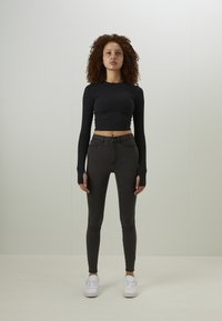 Svart, kortermet langermet topp i kombinasjon med mørke, tettsittende jeans. Modellen har på seg hvite joggesko, som fremhever den tettsittende stilen. Glatt stoffstruktur.