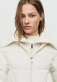 Joven mujer con cabello castaño claro usando una chaqueta acolchada blanca con dos cremalleras y un cuello alto frente a un fondo sencillo.