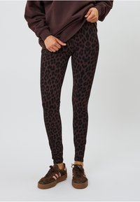Schwarz-braune Leo-Print Leggings mit figurbetontem Design, gefertigt aus einem glatten, dehnbaren Stoff. Getragen mit einem lockeren, dunklen Pullover und braunen Sneakers.