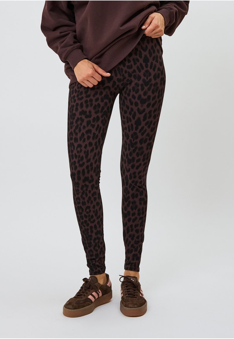 Schwarz-braune Leo-Print Leggings mit figurbetontem Design, gefertigt aus einem glatten, dehnbaren Stoff. Getragen mit einem lockeren, dunklen Pullover und braunen Sneakers.