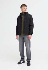 Must na zip-up jope kapuutsiga ja esipocketsitega, mille all on seljas roheline soonikkoeswiiter ja hallid teksad. Näha on valged triibud kolmes tulbas spordijalatsitel.