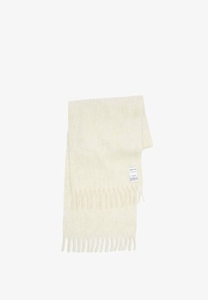 Écharpe blanche en tricot avec des franges, portée sur une chemise claire et un pantalon plissé noir. Des baskets vertes complètent la tenue.