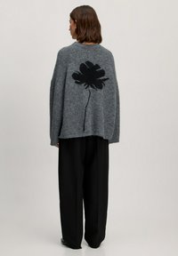Grauer Strickpullover mit lockerer Passform, der ein schwarzes Blumendesign auf dem Rücken hat. Kombiniert mit schwarzen weit geschnittenen Hosen für einen Kontrast.