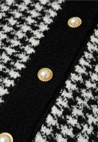 Tissu pied-de-poule noir et blanc avec une texture duveteuse, orné d'un bouton perle à accents dorés pour la fermeture sur une bande noire.