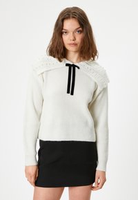 Weißer Pullover mit strukturiertem Strick, ausgestattet mit einem dekorativen Kragen mit Perlen und einem schwarzen Schleifenband. Kombiniert mit einem schwarzen Rock.