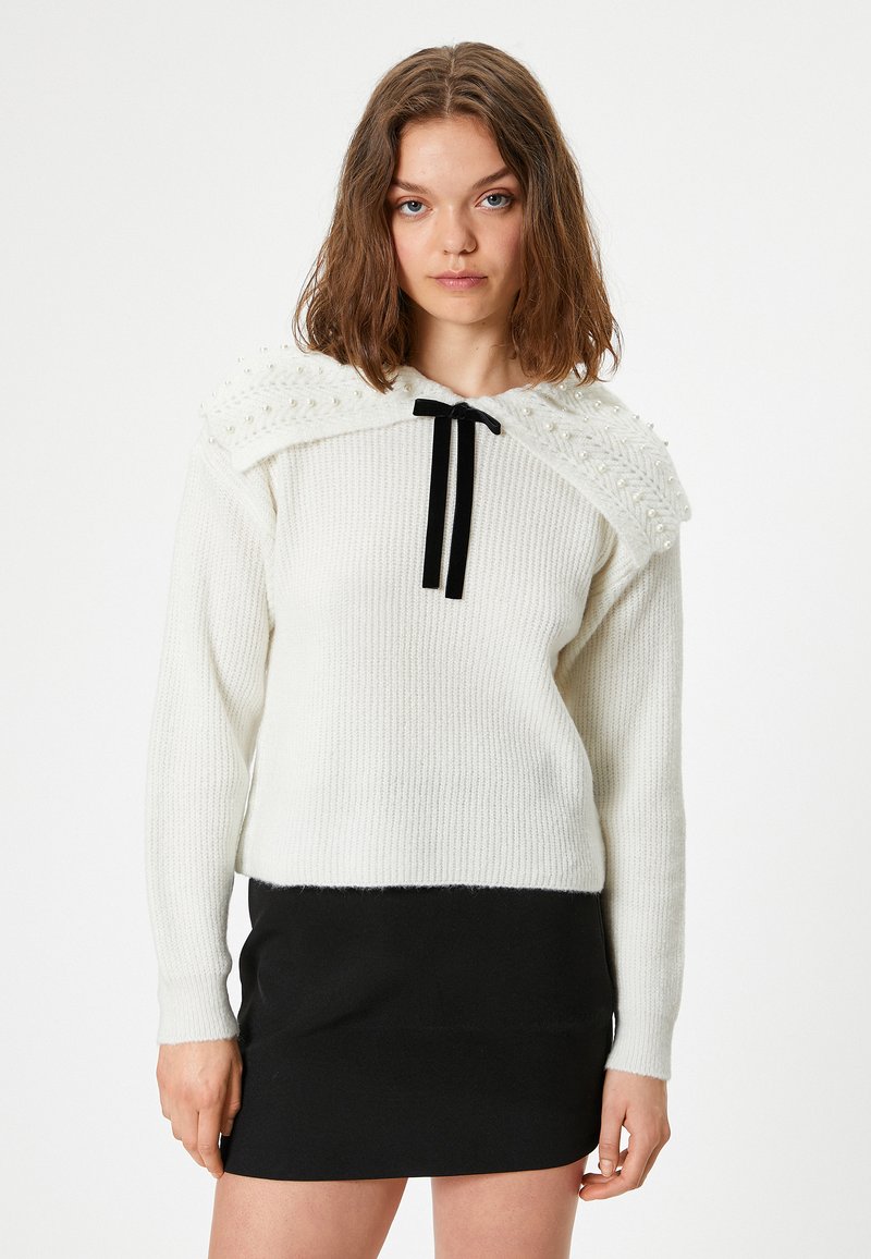 Weißer Pullover mit strukturiertem Strick, ausgestattet mit einem dekorativen Kragen mit Perlen und einem schwarzen Schleifenband. Kombiniert mit einem schwarzen Rock.