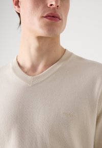 Beige v-hals genser laget av mykt strikket stoff, med ribbestrikket krage og subtile broderte logoer på brystet. Glatt tekstur.