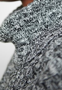 Strickpullover mit einem schwarz-weißen Muster, hohem Kragen und strukturiertem Stoff, der eine Mischung aus dicken und dünnen Garnen zeigt.