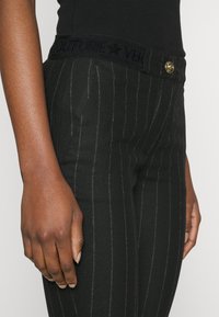 Close-up of a person wearing black pinstripe pants with a branded elastic waistband and gold button, paired with a black top.
