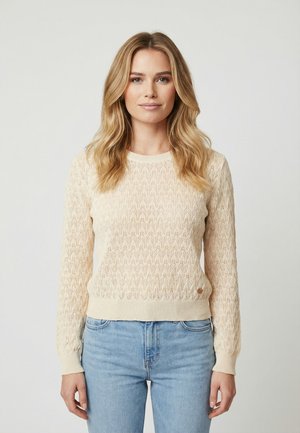 Mujer con cabello largo y rubio, vistiendo un suéter de punto color crema y jeans azul claro, de pie contra un fondo neutro y sencillo.