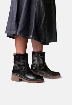 Jambes portant une jupe en jean foncé et des bottines noires brillantes avec fermeture éclair latérale et talons épais marron bas sur fond uni.