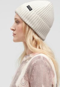 White ribbed knit beanie with a folded brim, featuring a black woven label. The model wears a pastel-patterned knit sweater.