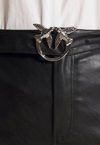 Ceinture en cuir noir avec une boucle circulaire en argent ornée de deux oiseaux se faisant face, présentant des détails et une texture raffinés.
