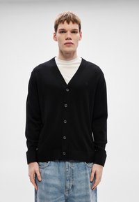 Cardigã preto feito de tecido macio com decote em V, abotoamento frontal e punhos canelados. Usado por cima de uma t-shirt branca e combinado com denim.