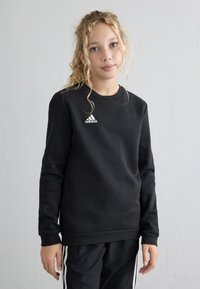 Giovane con capelli biondi ricci, vestito con una felpa nera Adidas e pantaloni neri con strisce bianche, in piedi davanti a un muro chiaro e semplice.