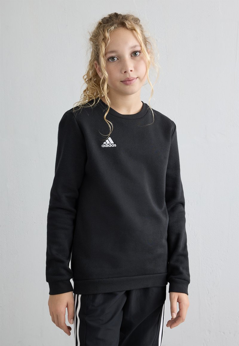 Giovane con capelli biondi ricci, vestito con una felpa nera Adidas e pantaloni neri con strisce bianche, in piedi davanti a un muro chiaro e semplice.