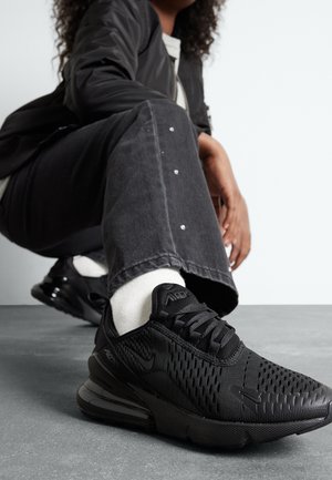 Person, der sidder på hug, iført sorte Nike Air Max-sneakers, hvide sokker, sorte bukser med nitter og en sort jakke mod en grå gulv og en hvid væg.