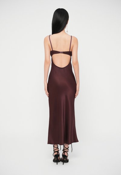 Žena s dlhými čiernymi vlasmi oblečená v bezrukávových hlboko hnedom saténovom šate s otvoreným chrbtom a čiernymi vysokými podpätkami s viazaním na šnúrky.
