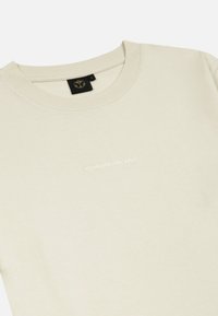 Lichtbeige t-shirt van katoen, met een ronde hals en een subtiele witte "TOMORROWLAND" logo op de voorkant. Het design is minimalistisch.