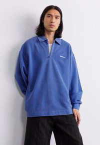 Blue pullover made of soft fabric, featuring a zippered collar, long sleeves, and a relaxed fit. Simple logo detail on the chest.