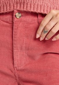 Roze corduroy broek met verticale ribbeltextuur, hoge taille en een knoopsluiting. Hand met een tatoeage rust dichtbij de tailleband.