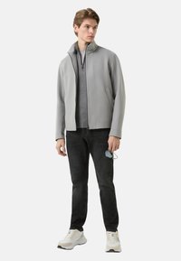 Lysegrå lynlåsjakke med høj krave over en grå skjorte, kombineret med sorte jeans og beige sneakers. Model holder solbriller.