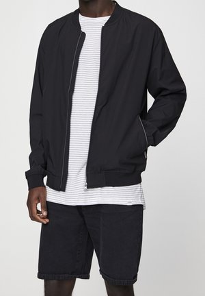 Man wearing a black zip-up jacket over a white and black striped t-shirt with black shorts against a plain background.