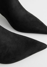 Botines de ante negros con un diseño de punta afilada. La superficie es suave con un acabado mate, mostrando una forma elegante y moderna.
