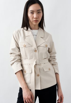 Kvinde iført en beige jakke med bælte og knappede bryst- og hofte lommer, over en hvid skjorte, stående mod en lys baggrund.