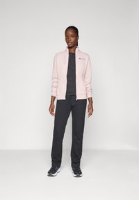 Veste zippée rose clair sur un T-shirt gris foncé, assortie à un pantalon de jogging gris foncé et des chaussures de sport noires. Design simple, sans motifs.