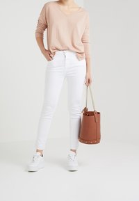 Jasnoróżowy sweter z długim rękawem i dekoltem w serek, zestawiony z białymi obcisłymi jeansami i białymi trampkami na platformie. Brązowa torba typu bucket z złotymi ćwiekami.