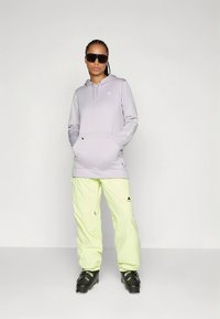 Világos lila kapucnis pulóver első zsebbel és logóval, világos zöld nadrággal és fekete csizmával párosítva. A modell napszemüveget visel, egyszerű háttér előtt áll.