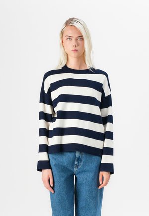 Maglione a righe blu navy e crema con collo a giro e maniche ampie, indossato con jeans in denim blu chiaro. Texture del tessuto a maglia liscia.