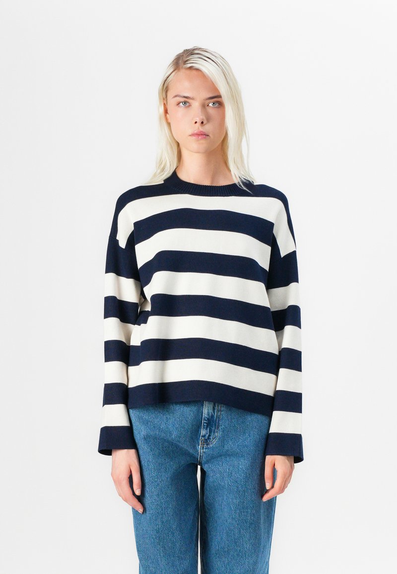 Pulover în dungi, cu nuanțe de bleumarin și crem, cu gât rotund și mâneci largi, purtat cu blugi din denim de culoare bleu deschis. Textură netedă, dintr-un material tricotat.