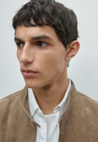 Beige Wildlederjacke über einem weißen Hemd mit Kragen, fokussiert auf die markanten Züge des Models und die kurzen, dunklen Haare vor einem neutralen Hintergrund.