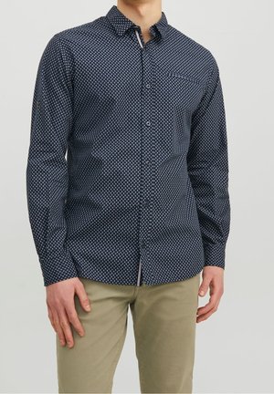 Homme portant une chemise bleu marine à manches longues avec de petits motifs blancs et un pantalon beige, debout devant un fond uni.