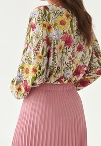 Blusa floreale con maniche lunghe di colore crema, caratterizzata da fiori rosa e gialli. Abbinata a una gonna rosa a pieghe. Tessuto testurizzato.