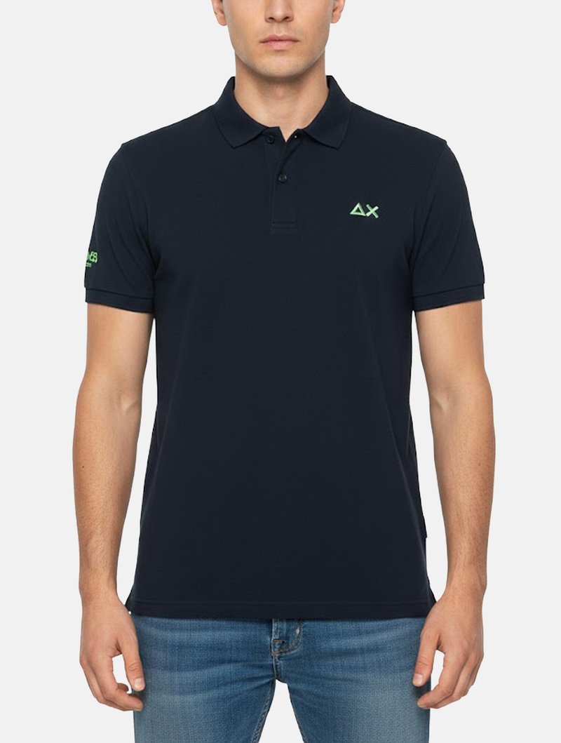 Uomo che indossa una polo aderente blu navy con logo triangolo verde e X sul petto a sinistra e testo verde sulla manica sinistra, abbinata a jeans blu.
