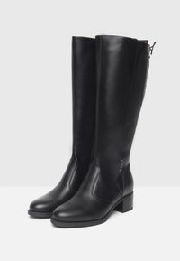 Une paire de bottes noires en cuir hautes avec des talons bas en bloc et un détail de laçage à l'arrière, présentées sur une surface blanche.