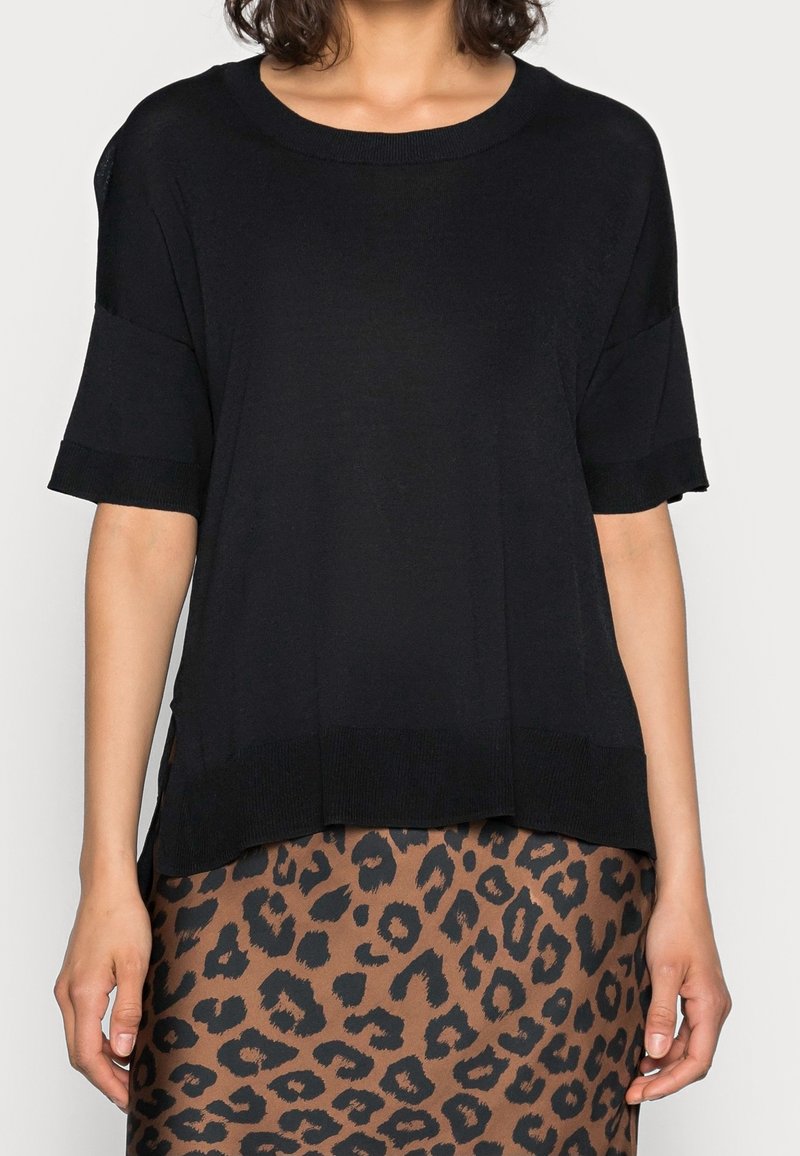 Kobieta ubrana w luźną czarną bluzkę z krótkim rękawem oraz brązowo-czarną spódnicę w cętki lamparta, stojąca na tle jednolitego tła.