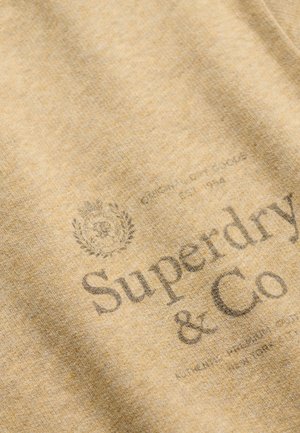 Tela marrón claro que muestra el logo descolorido de "Superdry & Co" con un pequeño escudo decorativo encima y texto adicional pequeño debajo.