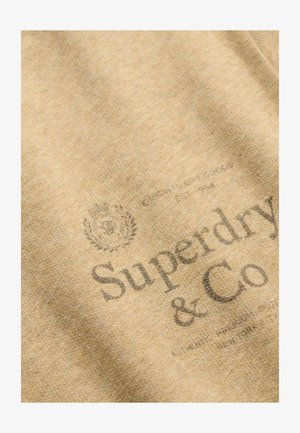 Tela marrón claro que muestra el logo descolorido de "Superdry & Co" con un pequeño escudo decorativo encima y texto adicional pequeño debajo.