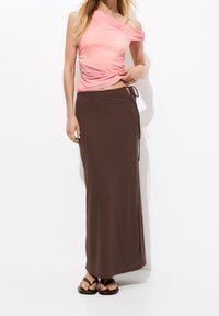 Haut rose à une épaule avec texture froncée, associé à une jupe portefeuille longue marron avec fermeture par nouage à la taille, porté avec des sandales plates.