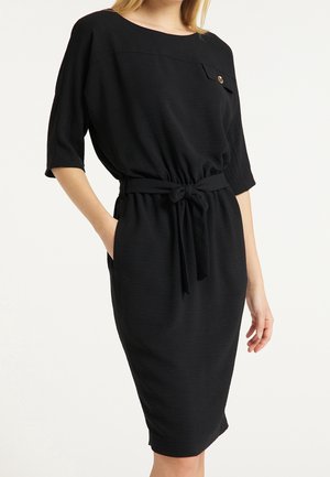 Femme portant une robe noire mi-longue avec des manches trois-quarts, une ceinture nouée à la taille et un bouton décoratif sur la poche poitrine.