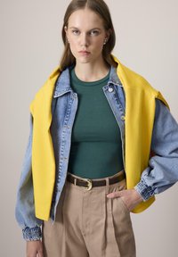 Sirge juustega naine, kellel on seljas pärliallased kõrvarõngad, roheline topp, teksajakk ja õlgadel kollane kampsun. Jalas on tal beežid püksid, millel on vöö, ja ta käsi on taskus.