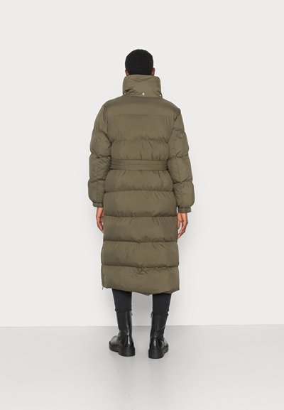 Pessoa vestindo um casaco puffer longo verde-oliva com gola alta, cintura cingida, calças pretas e botas ankle pretas, de costas para a câmara.