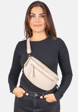 Frau trägt ein schwarzes Langarmshirt und dunkle Jeans mit einer beigen Umhängetasche, die auf ihrer Brust liegt, und hat die Hände in den Taschen.