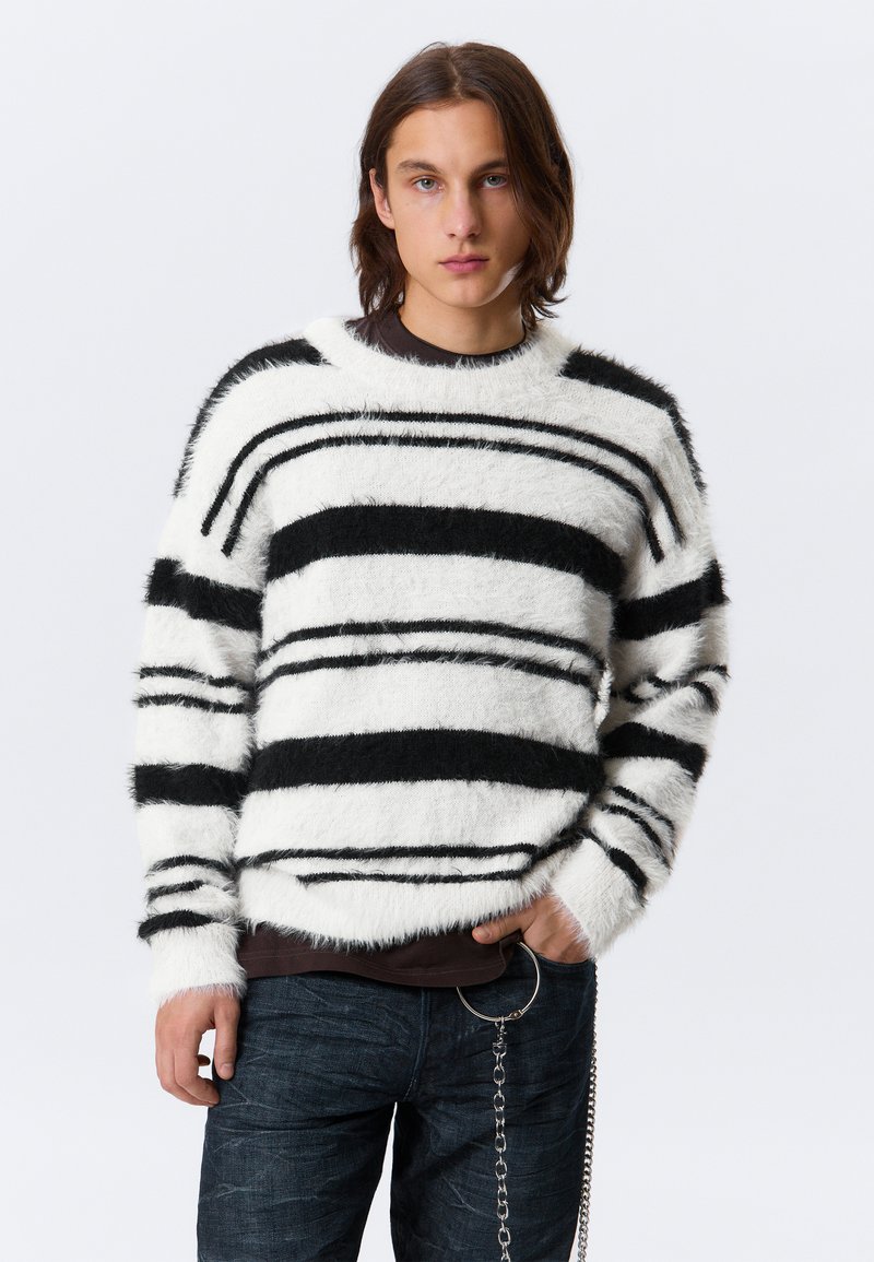 Jeune personne avec des cheveux bruns mi-longs portant un pull en peluche blanc avec des rayures noires et un jean noir avec un accessoire en chaîne.